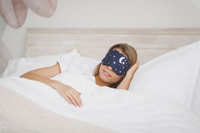 Jeune femme en t-shirt blanc, masque de sommeil, allongée dans son lit avec un drap blanc, une couverture et un oreiller, passant du temps dans sa chambre à la maison. Repos, détente, bonne humeur, concept de style de vie. Dormir avec la main sous la tête.