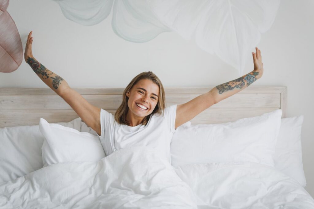 Jeune fille souriante en t-shirt blanc allongée dans son lit avec un drap blanc et une couverture d'oreiller, passant du temps dans sa chambre à la maison. Repos, détente, bonne humeur, concept de style de vie. Maquette de l'espace de copie. Etirer les mains en se levant.