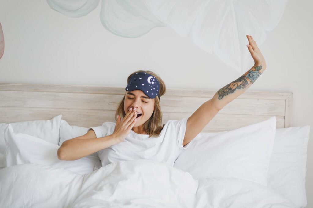 Jeune femme en t-shirt blanc, masque de sommeil, allongée dans son lit avec un drap blanc, une couverture et un oreiller, passant du temps dans sa chambre à la maison.